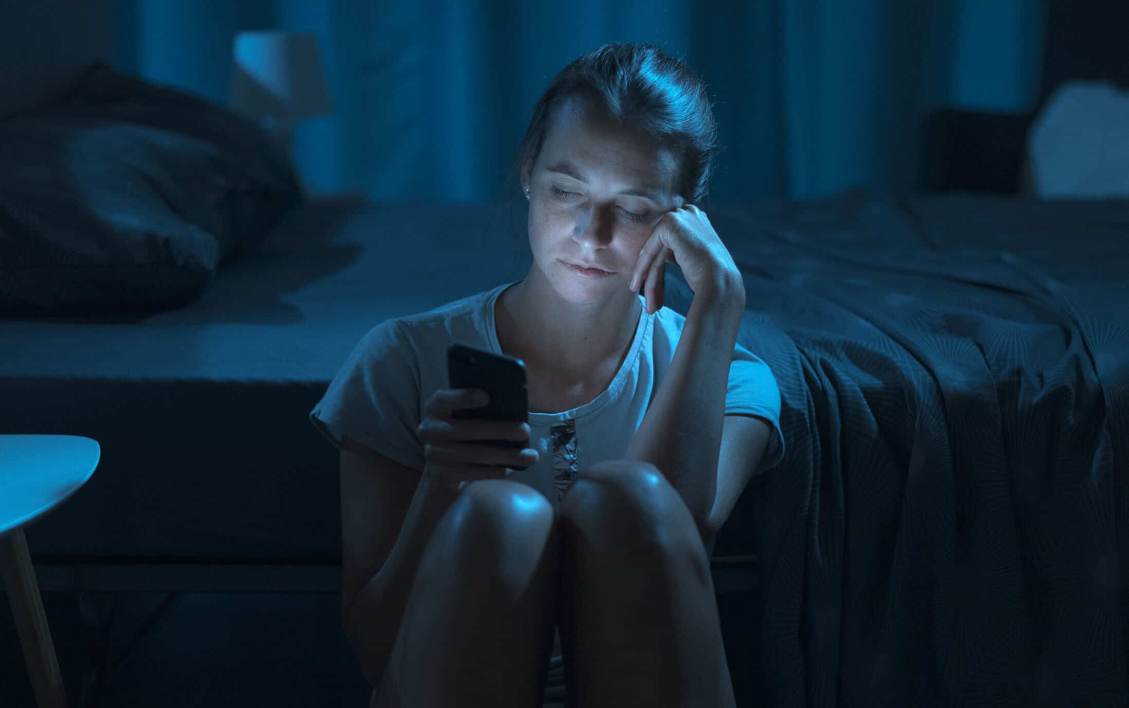 Schlaflose Frau mit Handy in der Hand im dunklen Zimmer sitzend auf dem Fußboden neben dem Bett.